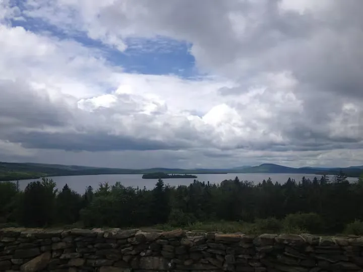 A lake with a small island, surrounded by trees and mountains under a cloudy sky.